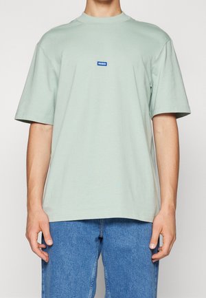 Mintgrünes Baumwoll-T-Shirt mit kurzen Ärmeln, Rundhalsausschnitt und einem blauen rechteckigen Logo auf der Brust. Trägt dazu eine blaue Jeans.