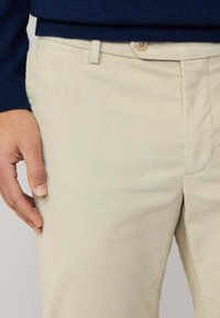 Pantalones de pana beige con una textura acanalada, que cuentan con un cierre de botón y bolsillos laterales. La cintura es lisa y tiene un lazo para cinturón.
