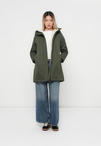 Parka imperméable verte avec capuche, dotée d'une douce doublure beige, de fermetures à boutons-pression et de poches latérales. Jean en denim clair à jambes larges et baskets noires.