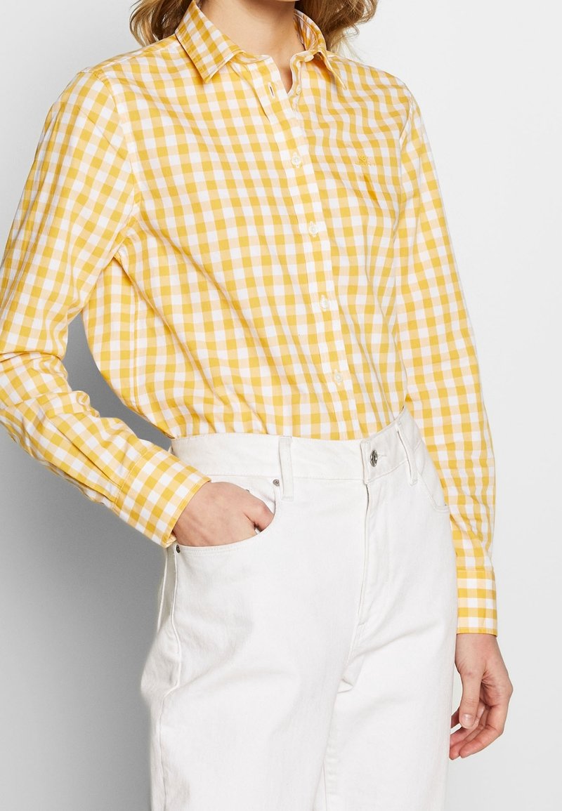 Femme portant une chemise à carreaux jaune et blanche boutonnée et rentrée dans un pantalon blanc taille haute, avec une main dans la poche.