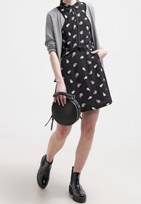 Schwarzes Kleid mit Paisleymuster, Knopfverschluss vorne und einem Gürtel, kombiniert mit einer grauen Strickjacke. Schwarze Ankle Boots und eine runde schwarze Handtasche.
