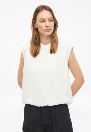 Vrouw met halflang lichtbruin haar, die een mouwloos wit topje en zwarte broek draagt, kijkt naar links tegen een witte achtergrond.