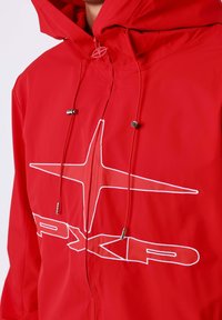 Rood gewatteerde jas met een wit omlijnd sterlogo en de letters "PXG" op de borst, voorzien van koordjes met metalen tips.