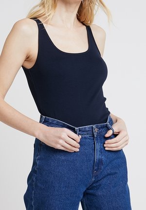 Vrouw draagt een nauwsluitend zwart tanktopje in een hoge taille blauwe jeans, met handen die de tailleband aanpassen.