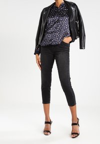 Giacca da moto in pelle nera sopra una blusa a fantasia blu navy, abbinata a jeans neri cropped e sandali con tacco neri.