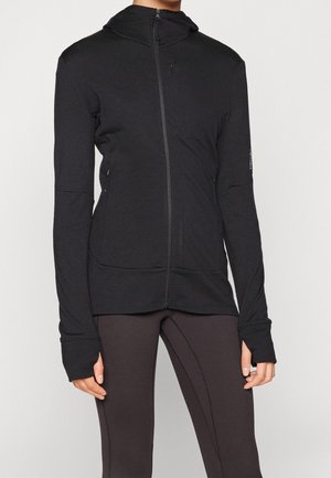 Kvinde iført en sort hættetrøje med lynlås og tommelhuller samt mørke tætsiddende leggings mod en ensfarvet baggrund.