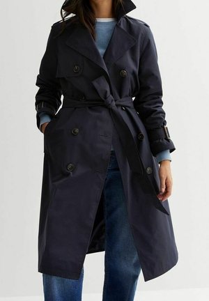 Trench-coat bleu marine à double boutonnage avec ceinture nouée à la taille, porté sur un pull bleu clair et un jean bleu, mains dans les poches.