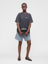 Γυναίκα που φοράει ένα σκουρόχρωμο oversized T-shirt, ανοιχτόχρωμα τζιν σορτς, μαύρα μυτερά φλατ παπούτσια, κρατώντας ένα μαύρο δερμάτινο τσαντάκι μπροστά με τα δύο χέρια.