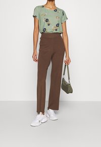 Zelená květinová blůza s krátkými rukávy, hnědé rozšířené kalhoty s žebrováním, bílé tenisky a malá olivová taška.