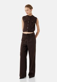 Top in maglia marrone tagliato, con maniche corte e bottoni frontali, abbinato a pantaloni larghi marroni a vita alta con cordoncino.