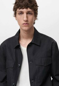 Zwarte jas met een kraag, voorzien van twee voorzakken en knoopsluiting. Draag boven een wit T-shirt. Zachte textuur, casual ontwerp.