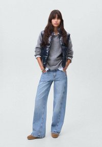 Marinblå pufferväst över en grå tröja, kombinerad med ljusblå vida jeans och bruna slip-on skor. Enkelt mönster och neutrala färger.