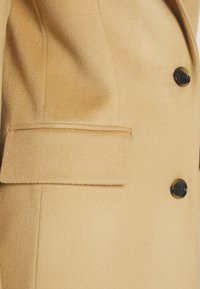 Cappotto beige in lana con una texture liscia, caratterizzato da una tasca a patta e due bottoni neri per la chiusura. Linee pulite e design su misura.