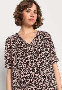 Blusa rosa com padrão de leopardos pretos, decote em V, mangas curtas com punhos dobrados e detalhes em botão na parte da frente.