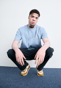 Light blue cotton t-shirt with short sleeves, yellow logo, black pants, and brown and white sneakers with green accents, against a neutral background.