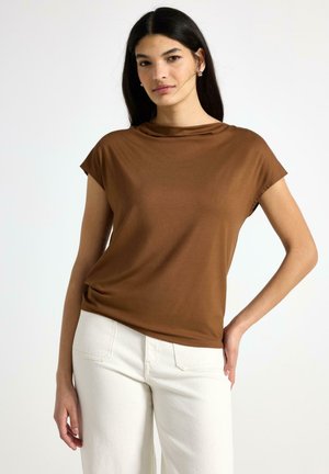 Kvinde med langt mørkt hår iført en brun kortærmet top og hvide bukser, stående med den ene hånd på hoften mod en hvid baggrund.