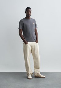 Tricou gri din bumbac cu un mic logo pe piept, asortat cu pantaloni largi bej și adidași bej cu accente maronii.