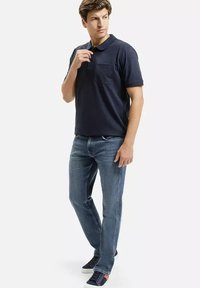 Granatowa koszulka polo z krótkim rękawem i kieszonką na piersi, w zestawie z niebieskimi dżinsami i ciemnymi sneakersami, stojąca na jasnym tle.