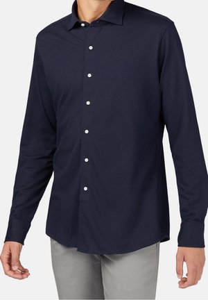 Mann trägt ein langärmliges, dunkelblaues Hemd mit Kragen und graue Hose, steht vor einem schlichten Hintergrund.