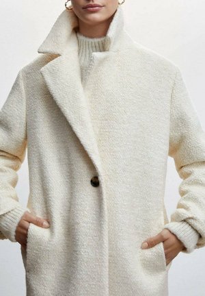 Donna che indossa un cappotto di lana color crema testurizzato con ampi risvolti e le mani in tasca, abbinato a un maglione a maglia coordinato.