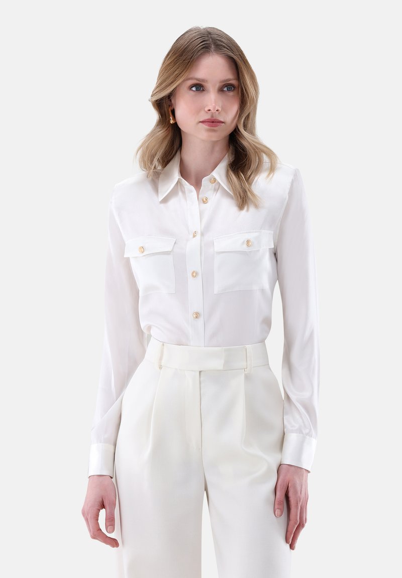 Camicia bianca a maniche lunghe in tessuto seta con chiusura a bottoni, dotata di due tasche sul petto e bottoni marrone chiaro.