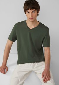 Olivengroen V-hals T-shirt van zachte stof, met korte mouwen, voorzien van een klein logo op de borst, gecombineerd met lichtbeige broek.