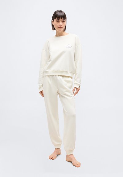 Femme debout pieds nus portant un sweat-shirt et un pantalon de survêtement assortis de couleur crème avec un petit logo sur la poitrine et la cuisse sur fond blanc.