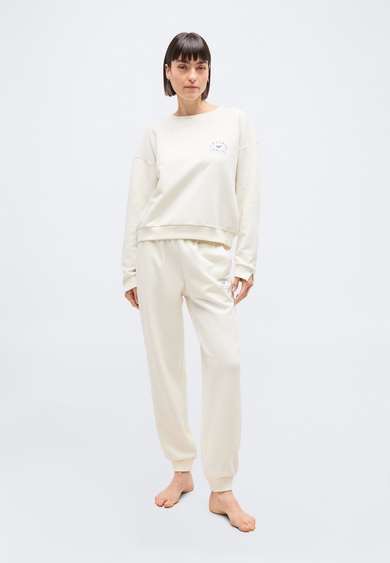 Γυναίκα όρθια, ξυπόλητη, φορώντας ασορτί εκρού φ ούτερ και παντελόνι φόρμας με μικρό λογότυπο στο στήθος και στο μηρό, σε λευκό φόντο.