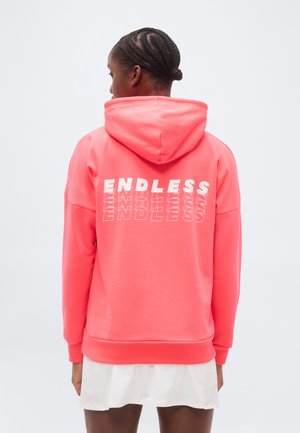 Femme aux cheveux tressés portant un sweat à capuche corail avec le mot "ENDLESS" répété en blanc au dos, associé à une jupe blanche.