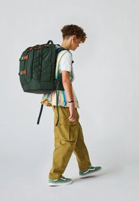 Grüner Rucksack mit braunen Lederakzenten, mehreren Außentaschen, gepolsterten Trägern und einem strukturierten Design. Wird von einer Person in Freizeitkleidung getragen.