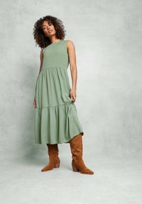 Grön, ärmlös midi-klänning med samlad midja och lager-på-lager kjol; kombinerad med höga, bruna mockastövlar med klack.