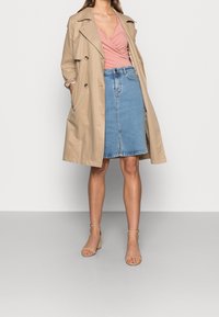 Donna che indossa un trench beige sopra un top avvolgente rosa, una gonna di jeans blu a lunghezza ginocchio e sandali beige con tacco a blocco, in piedi davanti a uno sfondo bianco.
