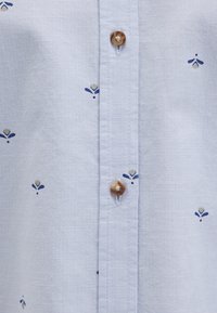 Light blue shirt featuring small embroidered floral patterns in navy, cream, and gold tones, with brown buttons along the front placket.