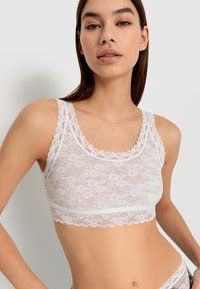 Bralette de encaje blanco con patrones florales, bordes festoneados y una textura suave. El diseño incluye tirantes anchos y una silueta ajustada.