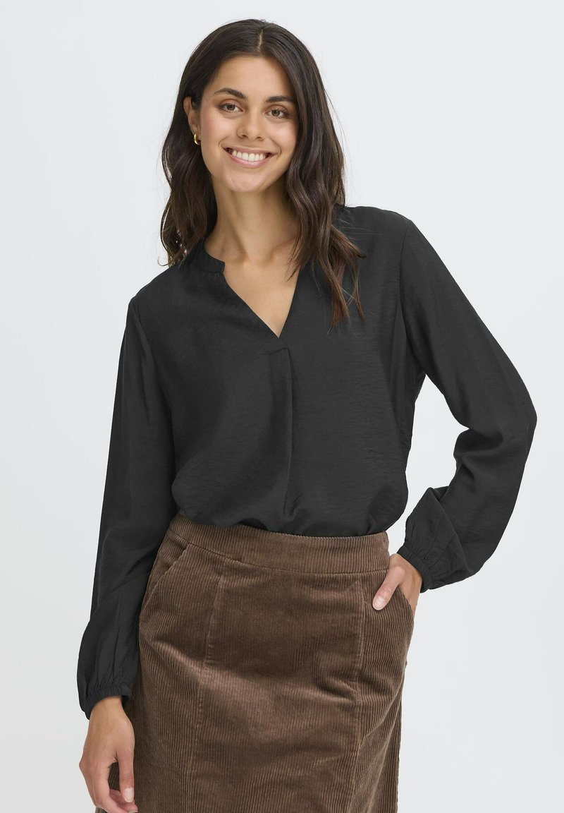 Young woman with long dark hair smiles, wearing a black long-sleeve blouse and brown corduroy skirt, hand in pocket.