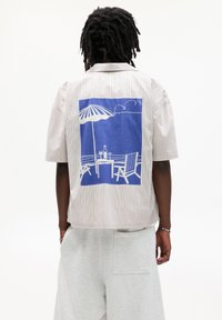 Camicia a maniche corte a righe in tonalità chiare con una grafica blu di una scena di spiaggia sul retro, che mostra un tavolo, un ombrellone e delle sedie.