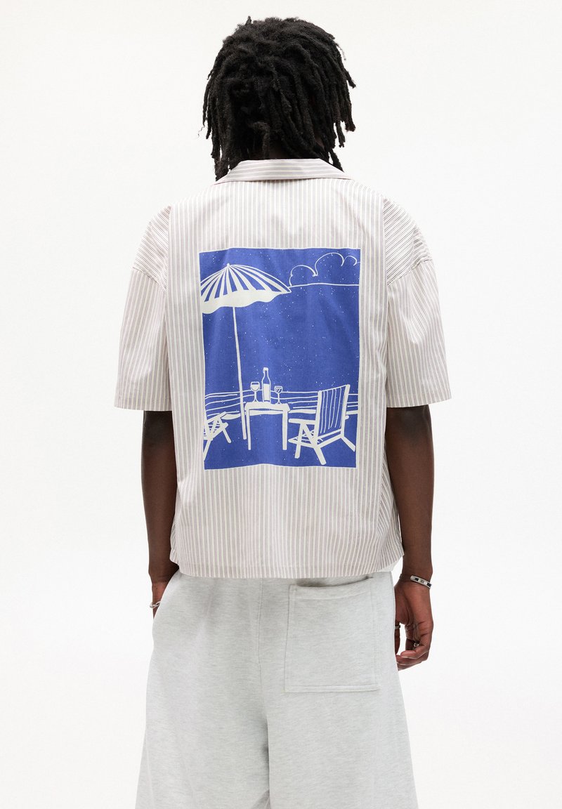 Camicia a maniche corte a righe in tonalità chiare con una grafica blu di una scena di spiaggia sul retro, che mostra un tavolo, un ombrellone e delle sedie.