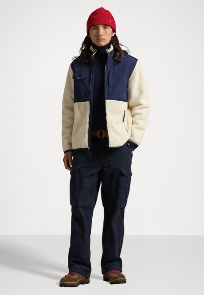 Fleecelineret jakke med marineblå og cremefarvede paneler, to brystlommer, parret med mørke cargo-bukser og brune snørebåndssko. Rød strikket hue.