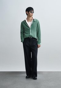 Πράσινη ριγωτή ζακέτα πάνω από λευκό πουκάμισο, σε συνδυασμό με μαύρο πλισέ παντελόνι και μαύρα παπούτσια· συνοδευόμενη από σκούρα γυαλιά ηλίου.