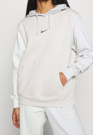 Kobieta ubrana w beżowo-białą bluzę z kapturem Nike z kieszenią z przodu, stojąca z jedną ręką w kieszeni na jednolitym tle.