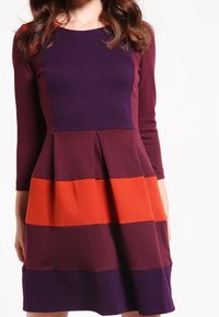 Sukienka z długim rękawem, charakteryzująca się poziomymi pasami w ciemnym purpurowym, burgundowym i pomarańczowym kolorze. Plisowany dół i dopasowana bodice o miękkiej teksturze.