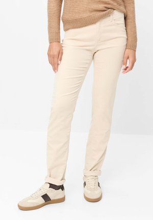 Person iført beige bukser, en brun sweater og lyse beige sneakers med mørkebrune accenter, stående på en hvid baggrund.