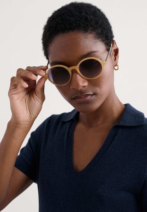 Occhiali da sole rotondi con una montatura traslucida color beige chiaro e lenti scure, indossati con una polo blu navy, tenuti da una mano.