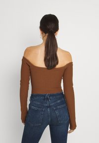 Hnědý žebrovaný off-shoulder top s dlouhými rukávy v kombinaci s vysokými tmavými džínami. Má upnutý střih a dvě zadní kapsy.
