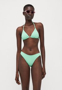 Bikini verde menta con tessuto testurizzato; parte superiore a triangolo con spalline sottili e parte inferiore a vita bassa; modella indossa occhiali da sole rosa.