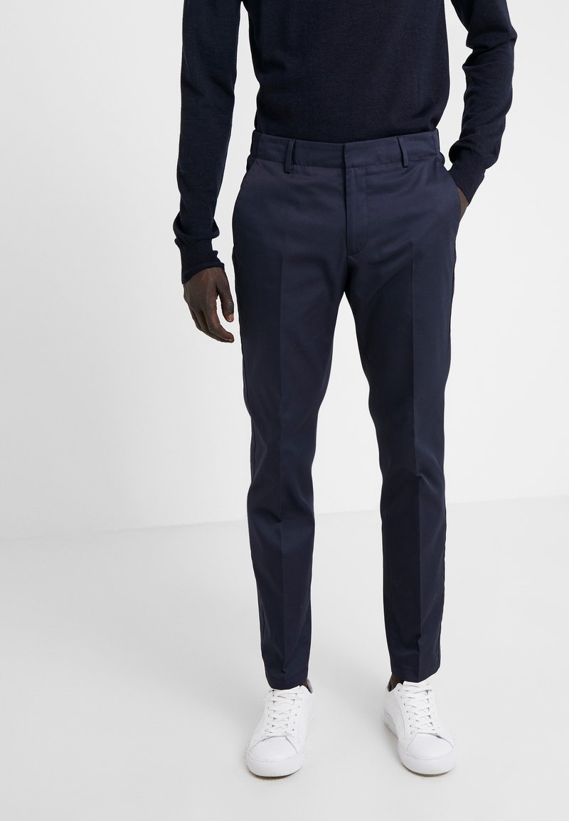 Námořnické kalhoty slim fit s hladkým povrchem a předními záhyby, v kombinaci s bílými teniskami. Oblečení doplňuje tmavý svetr.