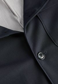 Gros plan d'un blazer noir montrant son revers, son bouton et sa doublure intérieure gris clair.