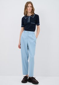 marineblaues Poloshirt mit hellblauer Kante und Tasche, kombiniert mit hellblauen hochgeschnittenen Hosen und dunkelbraunen klobigen Schnürschuhen.