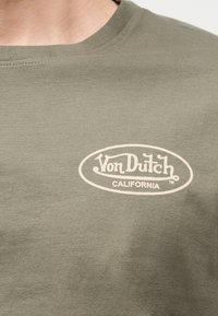 Κοντινό πλάνο ενός πράσινου μπλουζάκι με μπεζ λογότυπο «Von Dutch California» τυπωμένο στο πάνω μέρος του στήθους.