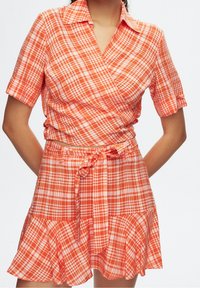 Orange ruta kort klänning med omslag, korta ärmar, knapp-up krage, midjeband och en utsvängd volangnederdel. Tillverkad av bomull.